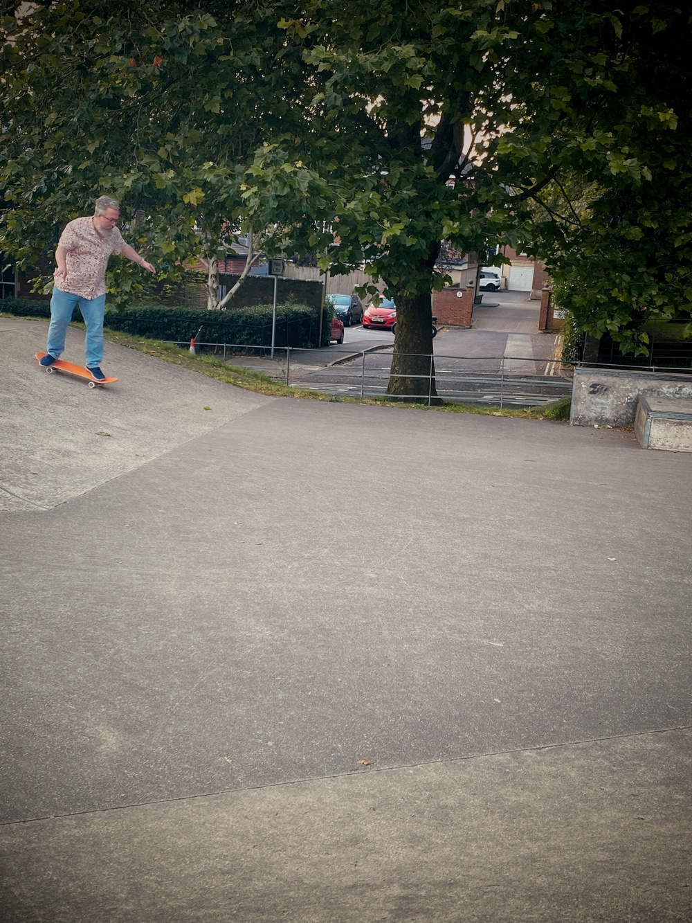 A person skateboarding down a ramp in an outdoor skatepark surrounded by trees. In the background, there are parked cars and a residential area. The person is wearing a patterned shirt and jeans.