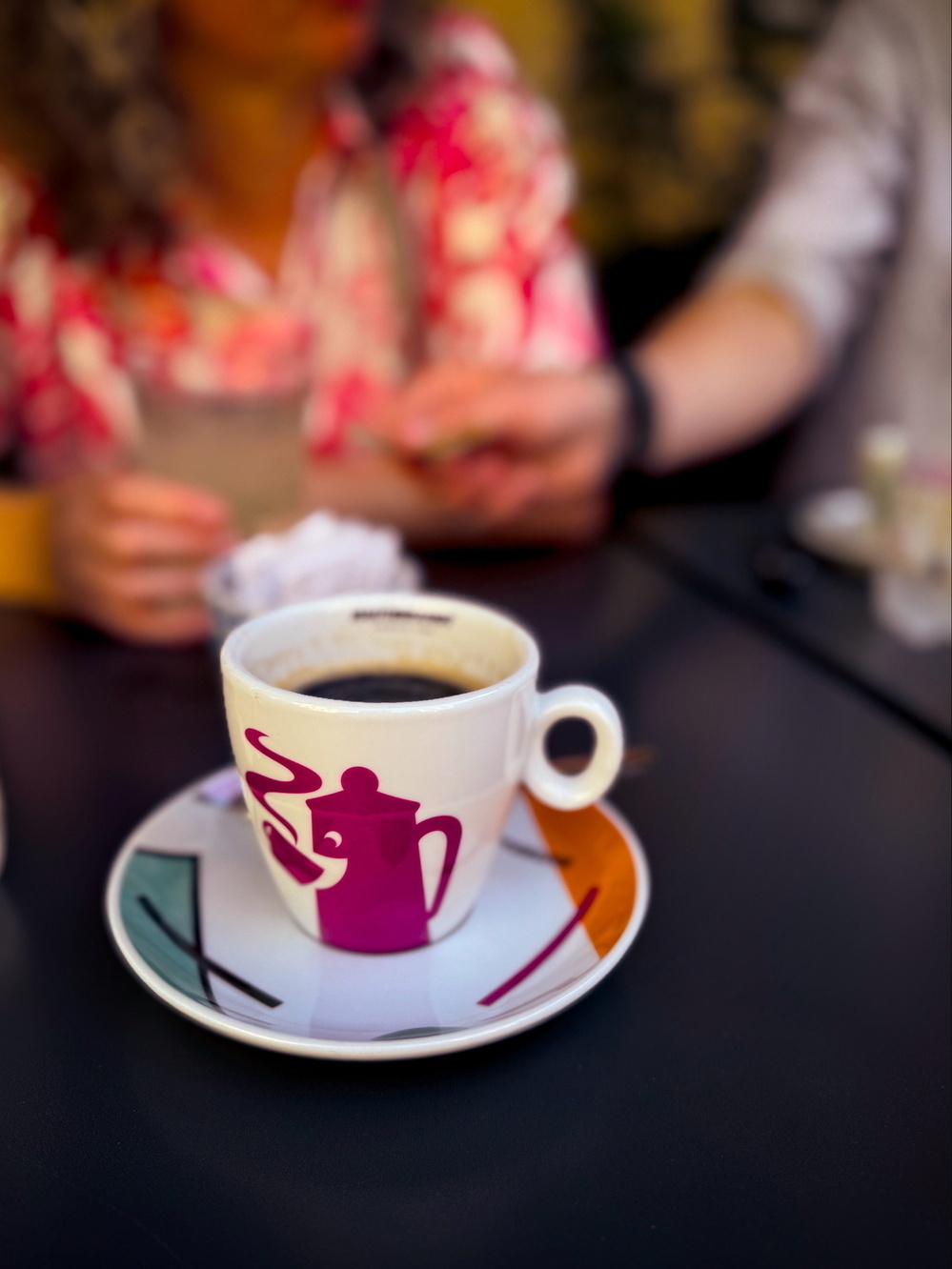 A cup of coffee on a colorful saucer featuring a graphic of a coffee pot, placed on a dark table. In the background, two people are sitting and one is wearing a bright floral shirt, but they are out of focus.