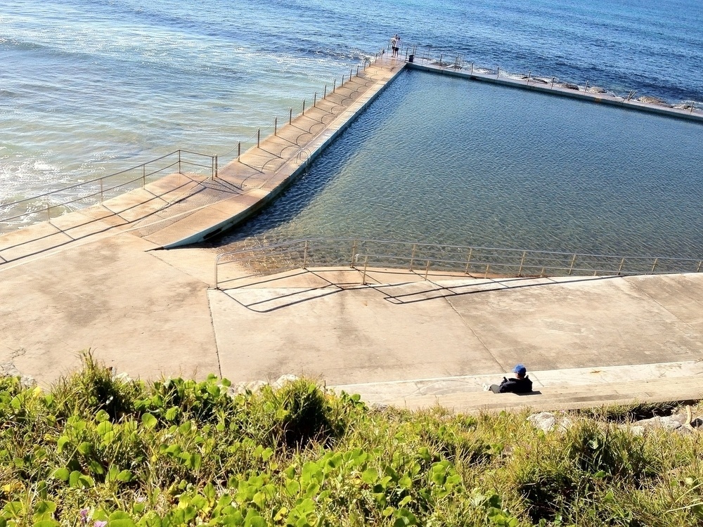 A sunny but cold day, looking down from a clifftop crowned with grass and nasturtium leaves, at an empty beach swimming pool. The surface of the pool is rippled but flat. We can see half the pool and two of its concrete walls, lined with chains and bollards, jutting out at an angle into the sea. Two people stand together at the very furthest point. Below the cliff is a set of steps running the length of the pool, and a broad concrete walkway at the pool’s edge, with a ramp and railings descending to the water. A man with a phone sits alone in the middle of the steps, dressed in trainers and dark pants, dark jacket with wide collar, blue cap and glasses. We see him from above. In both the ocean and the pool, the water is deep blue, shading to green in the shallows.