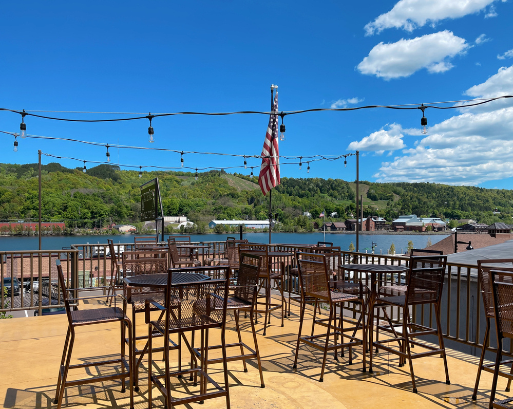 Looking at the Keweenaw waterway from a brewery terrace with empty chairs and tables, and a US flag in the corner in front of the green hills