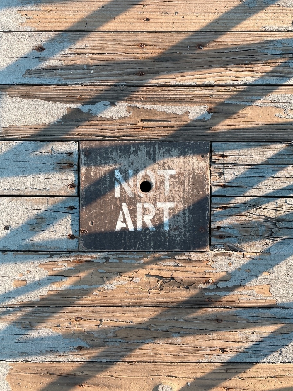 A utility cover in a wooden walkway reading "NOT ART", with crossing shadows