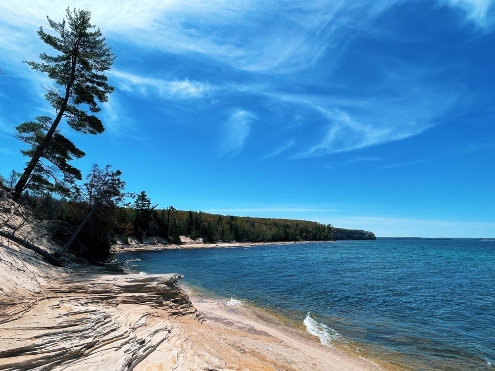 A view of the lake superior shoreline featuring a warped tree, swirling rocks, and whispy clouds