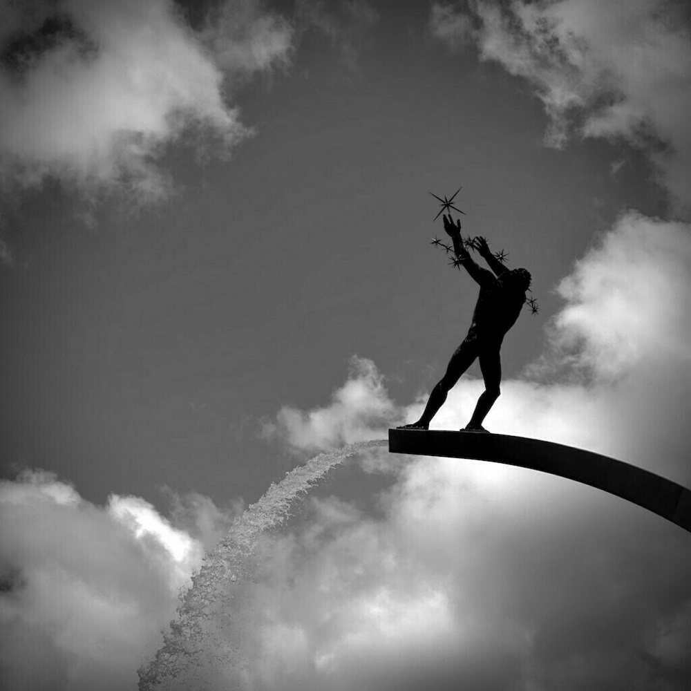 Black and White statue of figure with stars looking up at clouded sky. Water spewing from statue. 