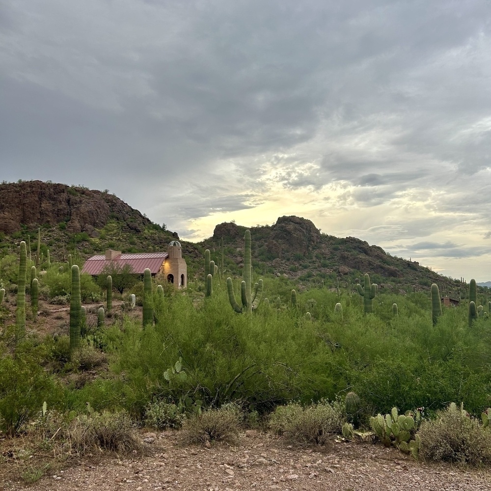 A simple chapel with a red roof stands among saguaros and lush greenery in a rugged desert landscape and under a cloudy sky.