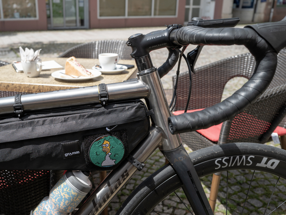 Upper front triangle of a titanium road bike with a carbon fork and handlebar. Focus is on a velcro patch on a black frame bag showing the Homer Simpson disappearing into a hedge meme. In the background a table with coffee and cake is visible, slightly blurred. 