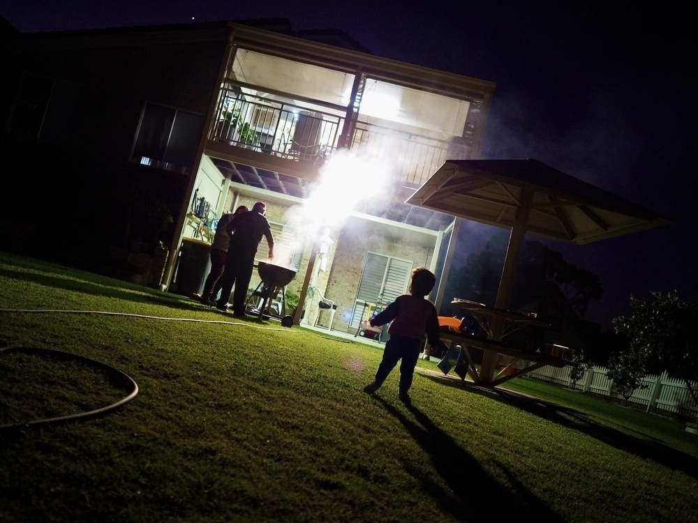 Set in a backyard, a toddler looks at an illuminated house with two men barbecuing in a big cloud of smoke.