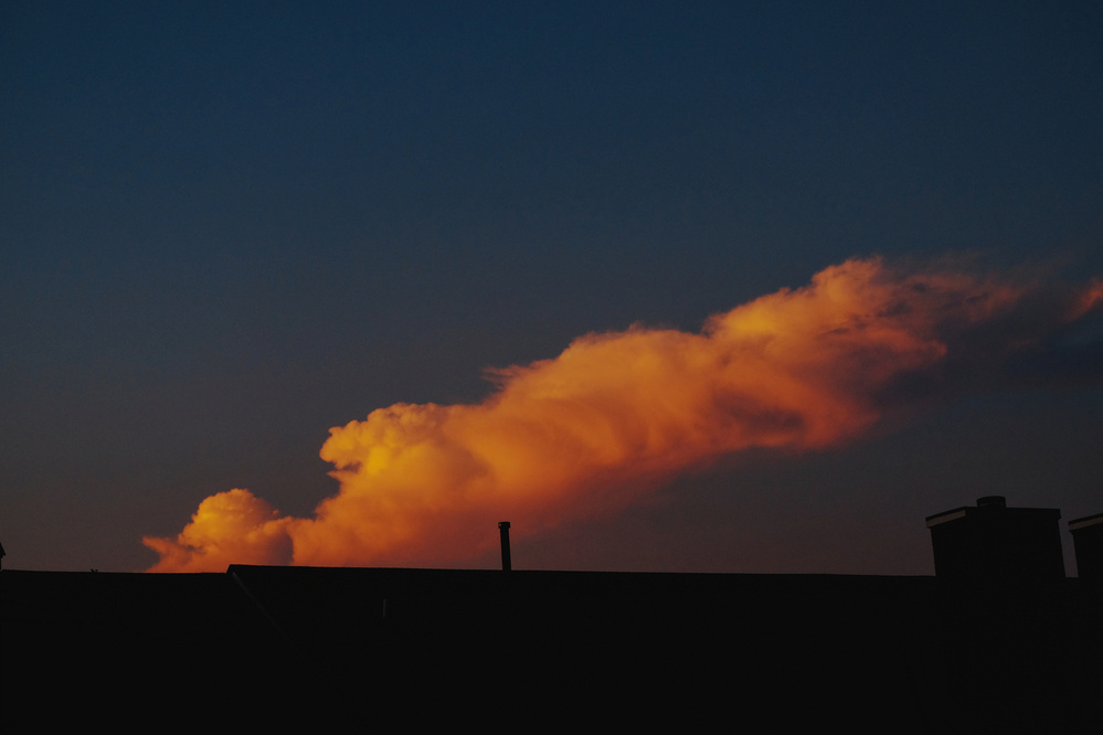 Silhouette of a roofline with a long and fluffy pink, red, and yellow cloud against the darkening sky.