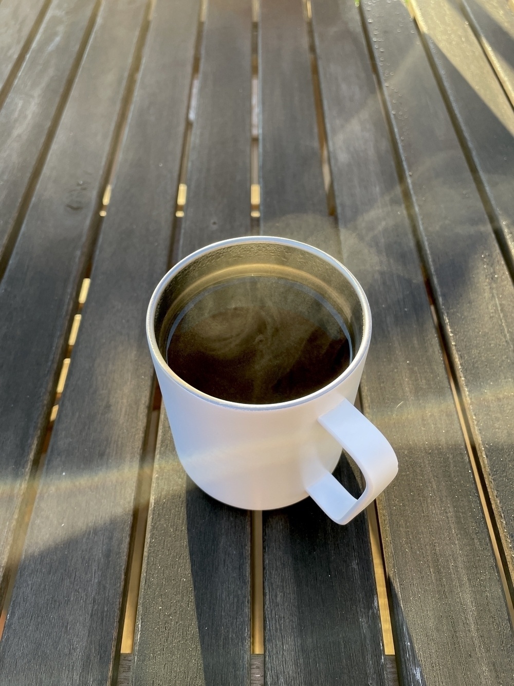 Early morning sun splashes over a wooden table top with a white metal mug of steaming coffee in the center