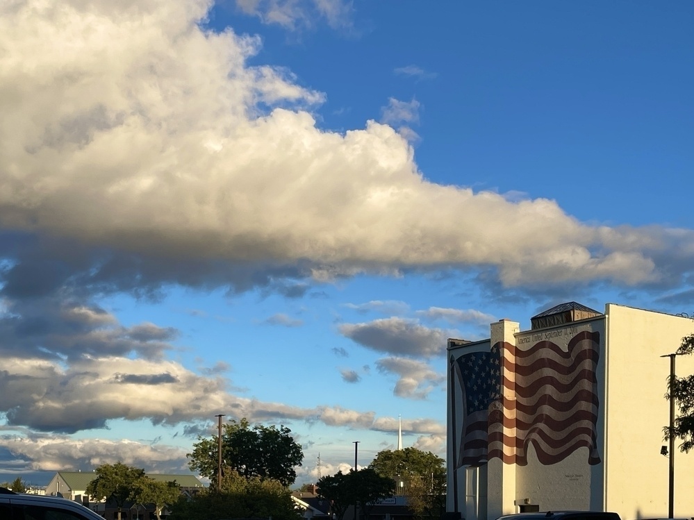 Large Fluffy clouds over a blue sky with sunset yellowing a plain block building in the foreground with an American flag painted across it