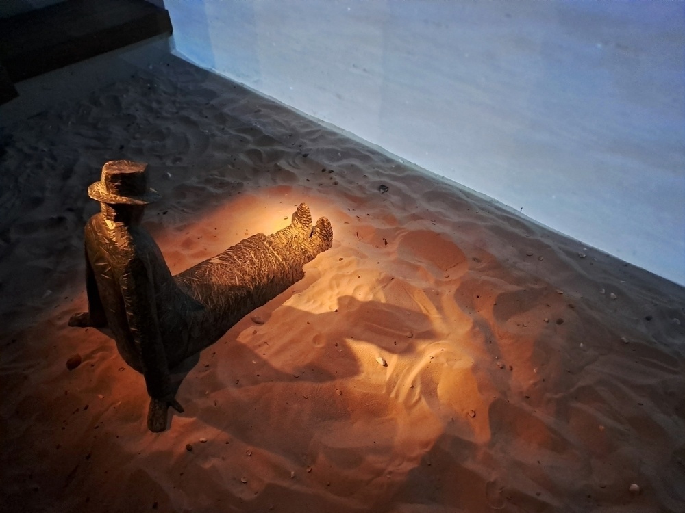 standbeeld van een zittende man in zand  kijkend naar een scherm waar de zee op getoond wordt
