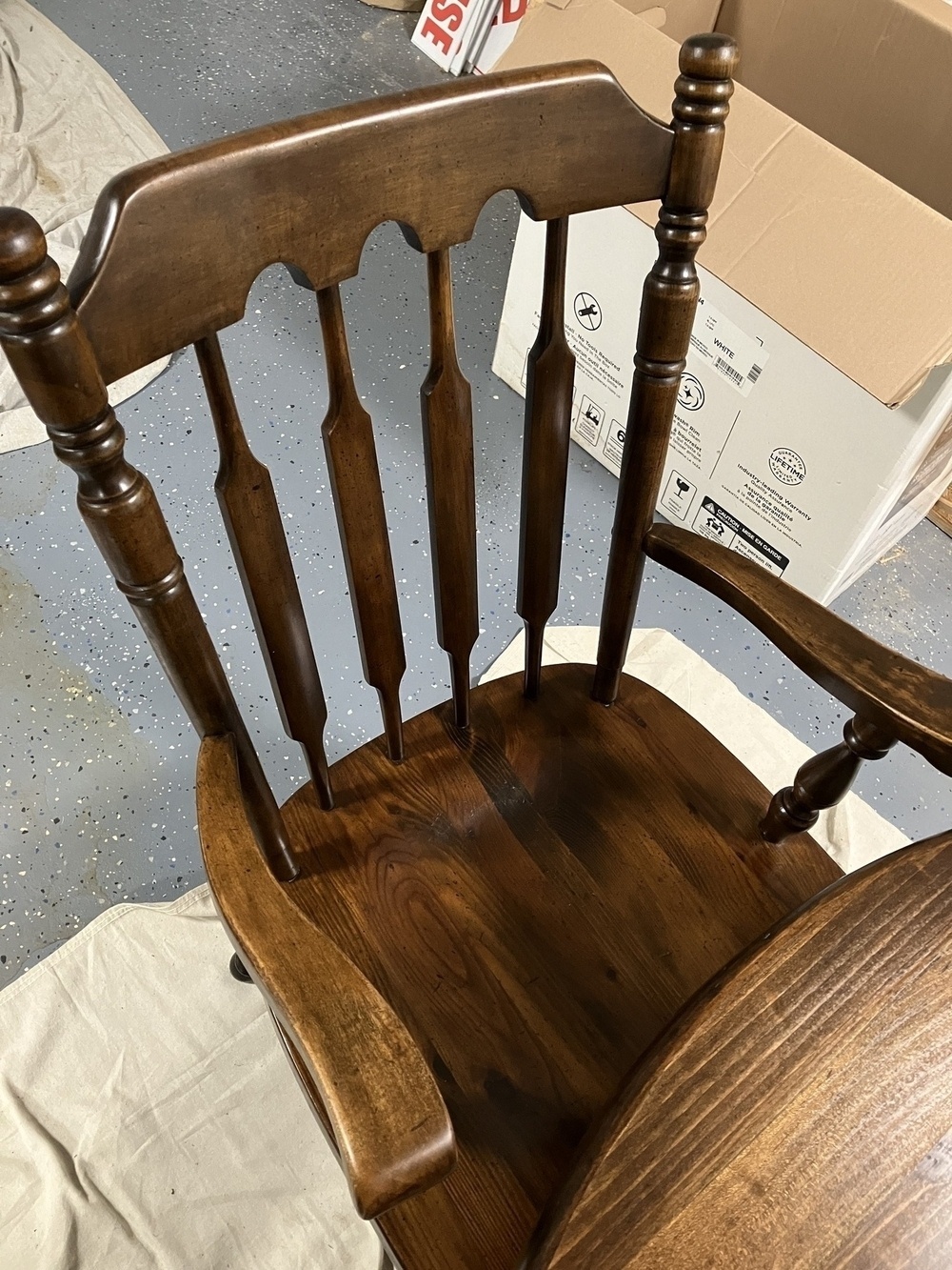 Auto-generated description: A wooden chair with a spindle back design is positioned on a white cloth with a cardboard box and other items in the background.