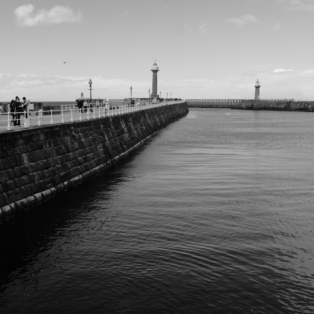 A black-and-white photograph features a long pier with railings, people walking along it, and lighthouses at both ends, extending out into a calm body of water under a partly cloudy sky.