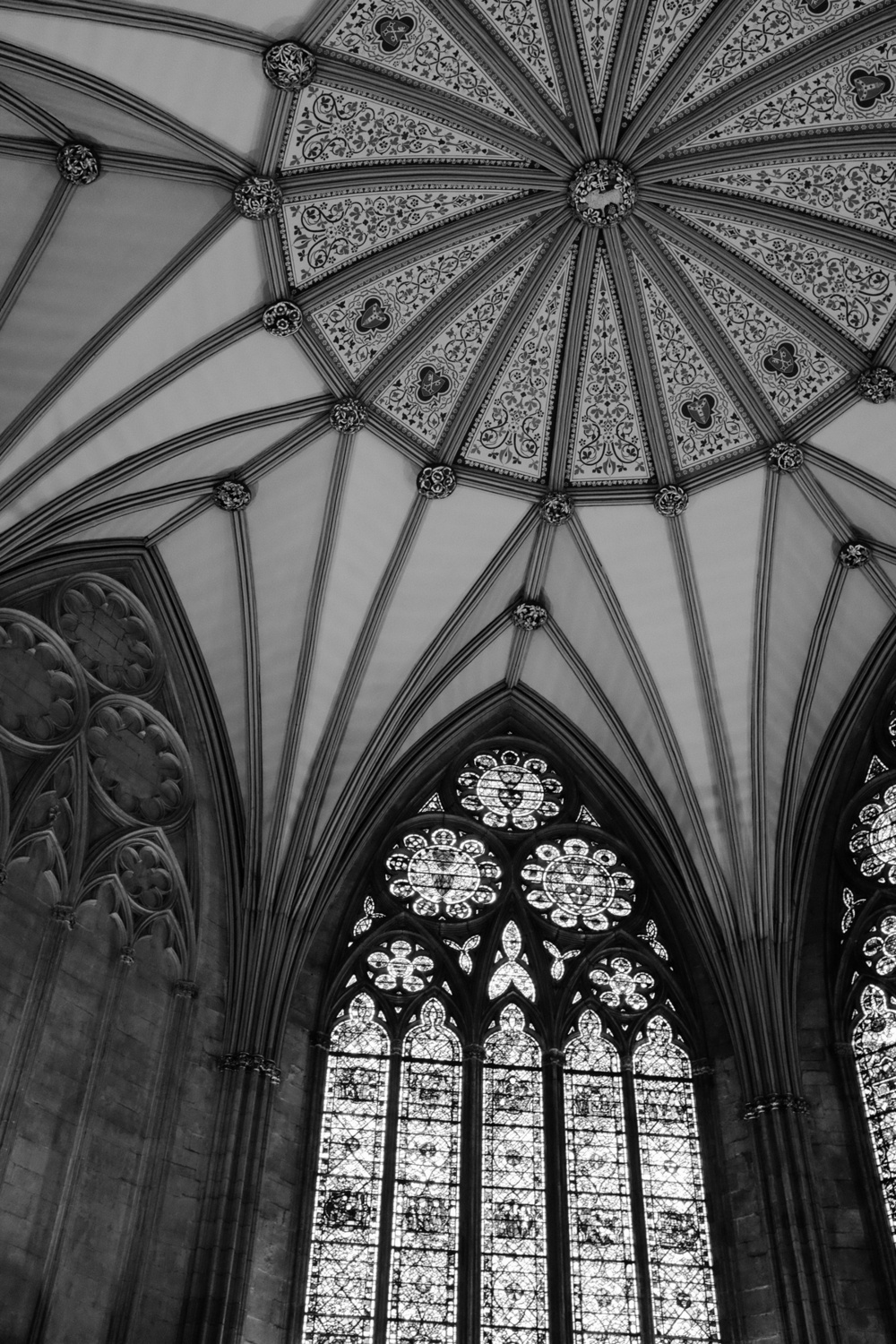 The top arch of a stained glass window points up to the central point of a high, vaulted ceiling, in black and white.