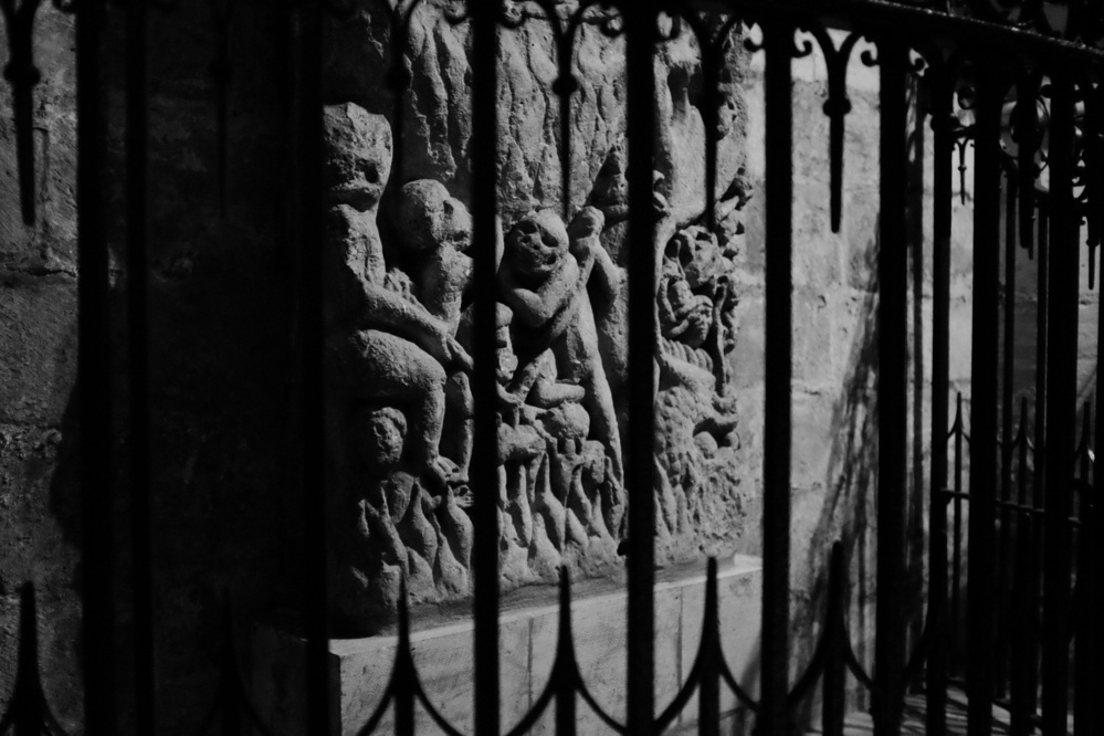 Creepy demonic-esque figures carved into a stone relief, viewed through an iron grate in black and white.