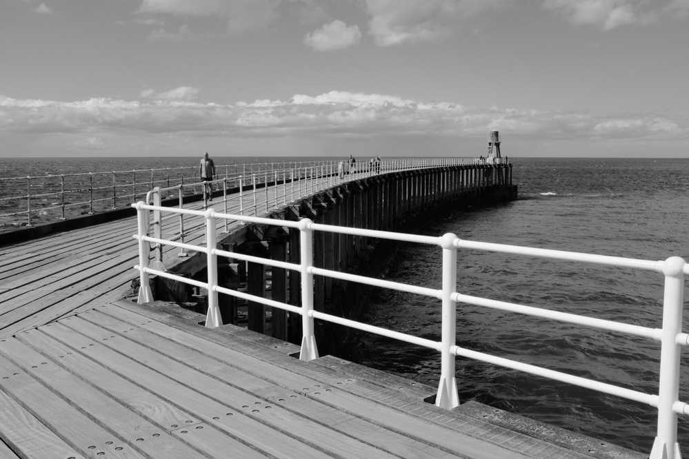 A long pier extends out into the ocean under a partly cloudy sky, with a few people walking along it.