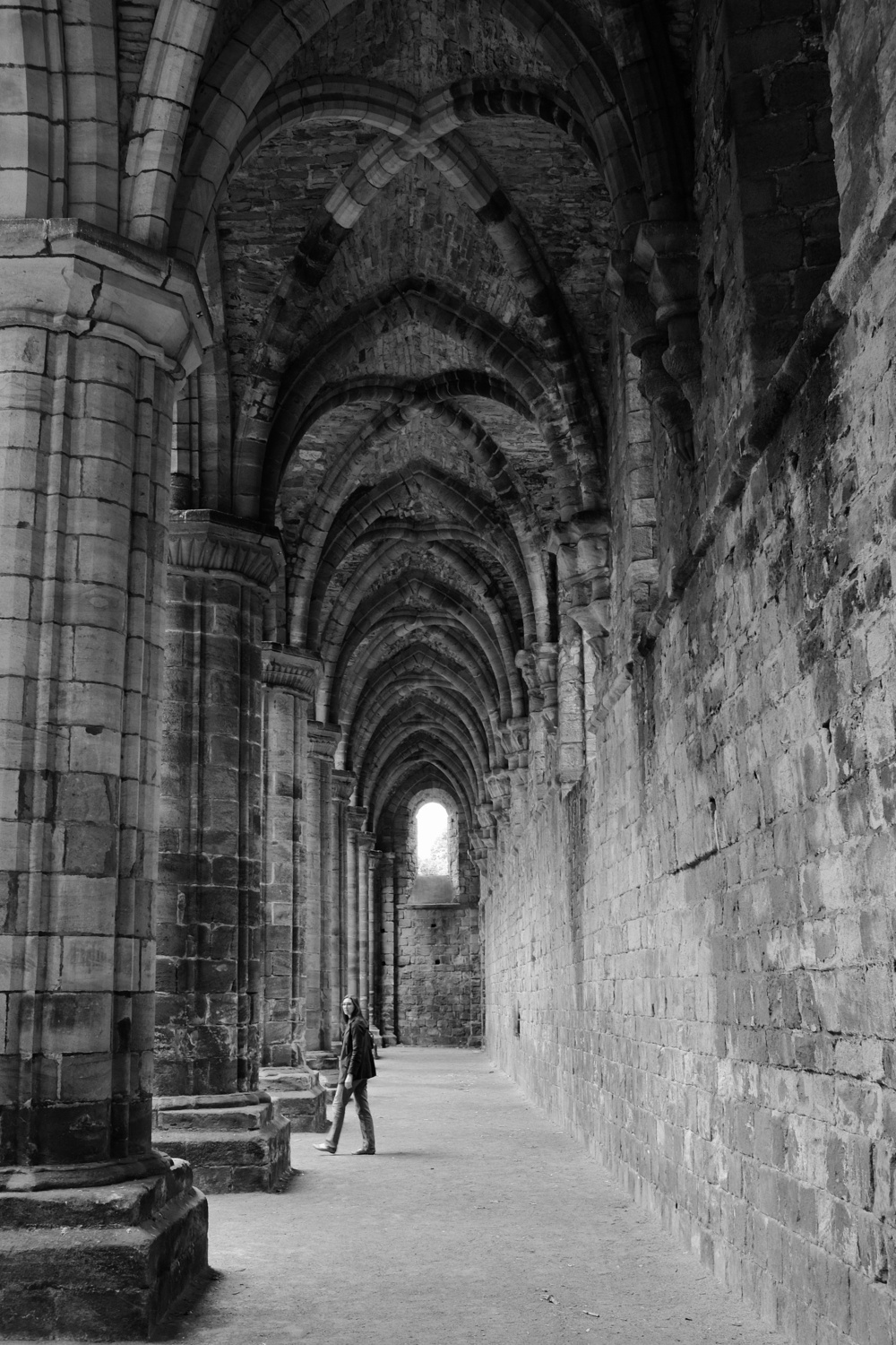 A long hallway with tall stone archways, Jacqueline turns to the left to walk between two stone columns