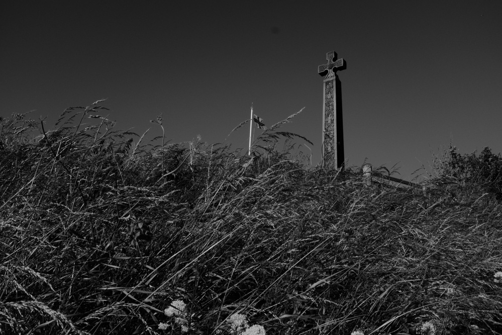 A stone cross monument stands among tall grasses, with a flag in the background under a clear sky.