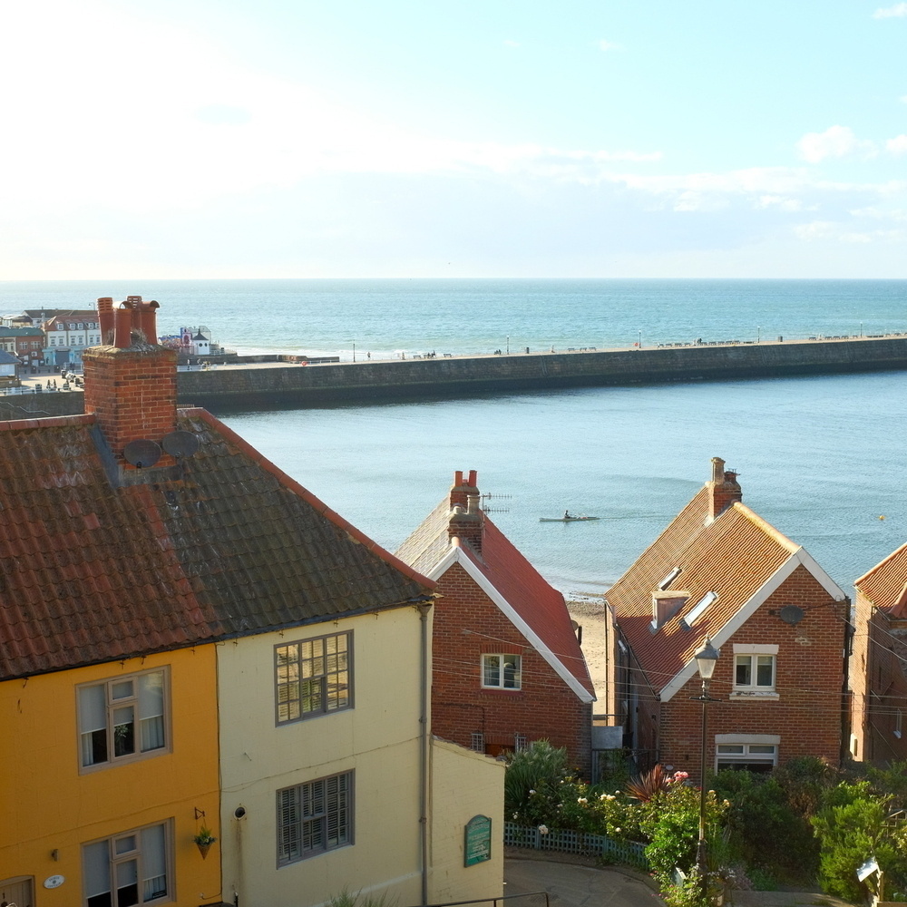 A coastal town features charming houses with red roofs overlooking a scenic ocean view with a long pier extending into the water.