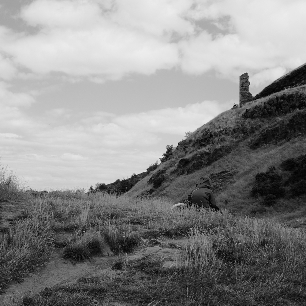 A hiker sits down to rest, with a view of the St. Anthony Chapel ruins on a hill in the distance. Black and white.