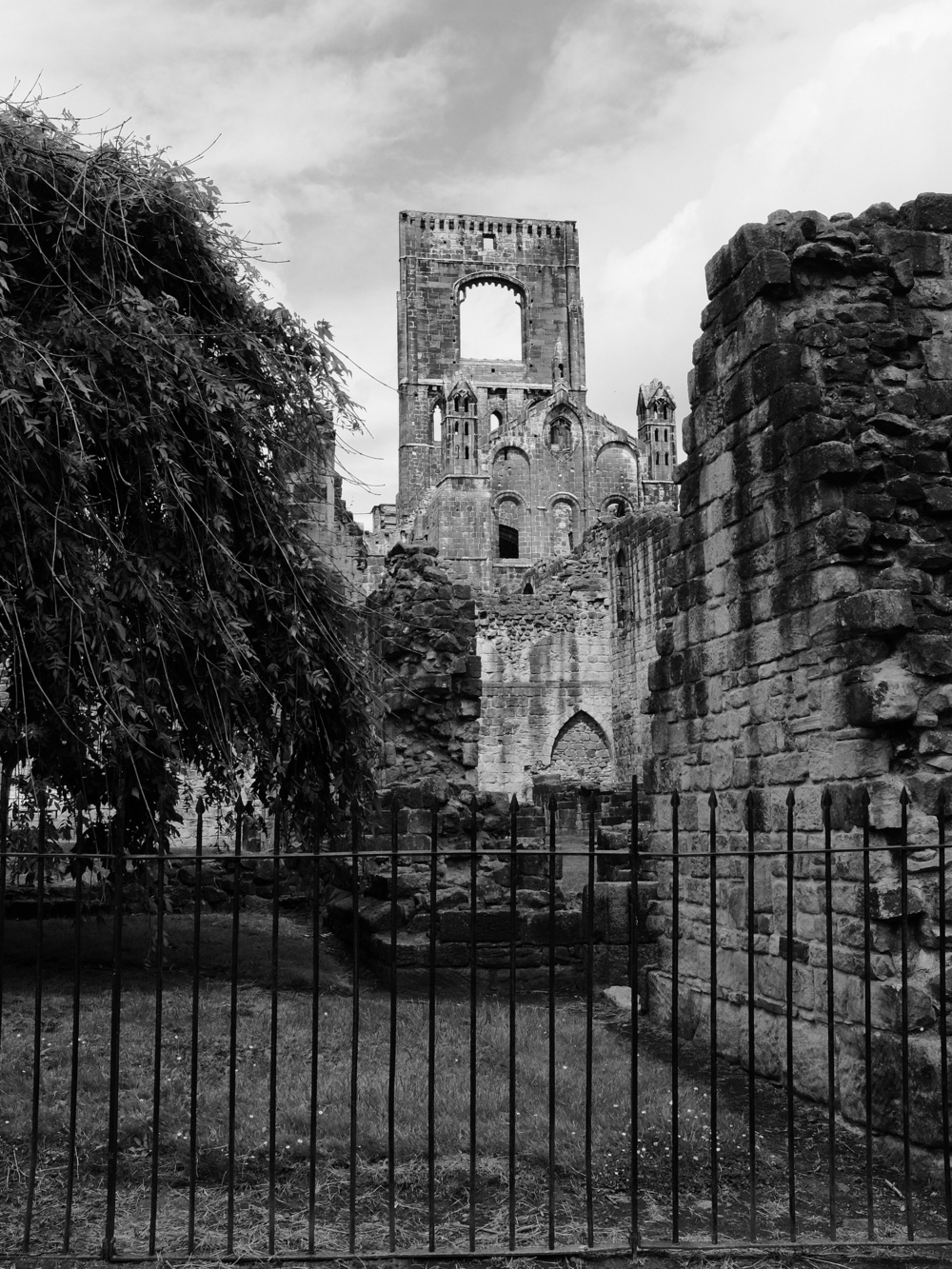 The ruined walls of the monastery arise in the background, with trees and a dilapidated brick wall in the foreground and an iron-wrought fence in the foreground