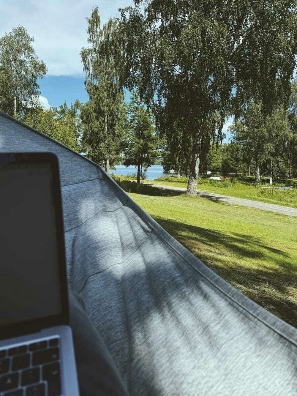 View from a hammock overlooking trees and a lake with a glimpse of a laptop on a pillow in the foreground.