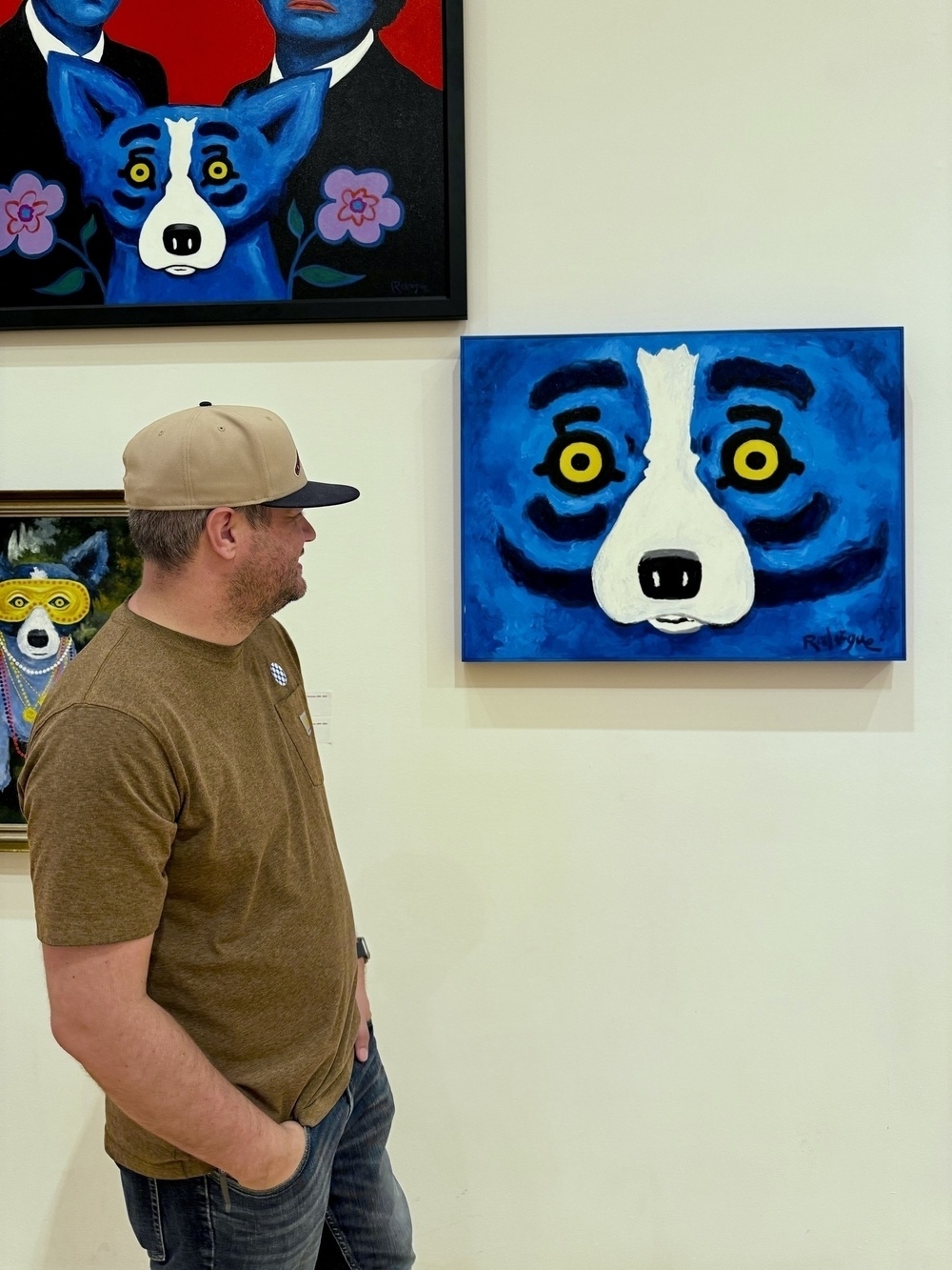 A man wearing a tan cap and brown shirt is looking at a painting of a blue dog with yellow eyes in an art gallery.