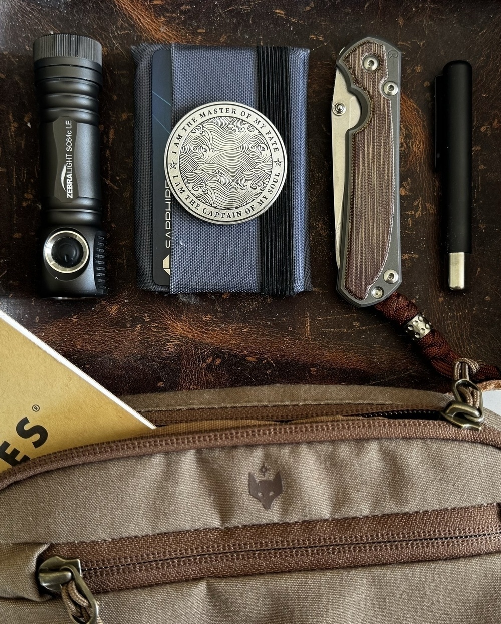 Flashlight, knife, coin on a wallet, and pen neatly arranged on a leather surface; tan bag with notebook partially visible. Coin text: “I AM THE MASTER OF MY FATE, I AM THE CAPTAIN OF MY SOUL.”