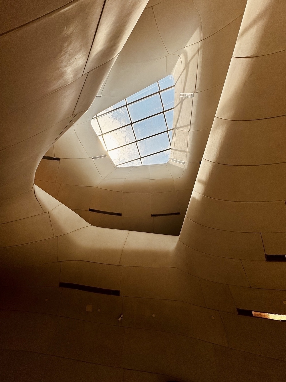 Skylight window illuminates undulating beige walls and ceiling, blending organic curves and sharp lines, with scattered square and round vents, creating a futuristic architectural interior under a blue sky.