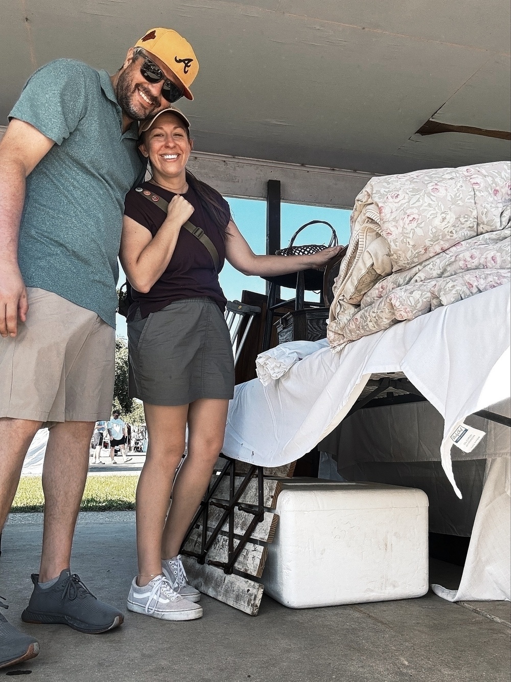 A smiling man and woman stand outdoors next to a table covered with blankets and other items, under a canopy.