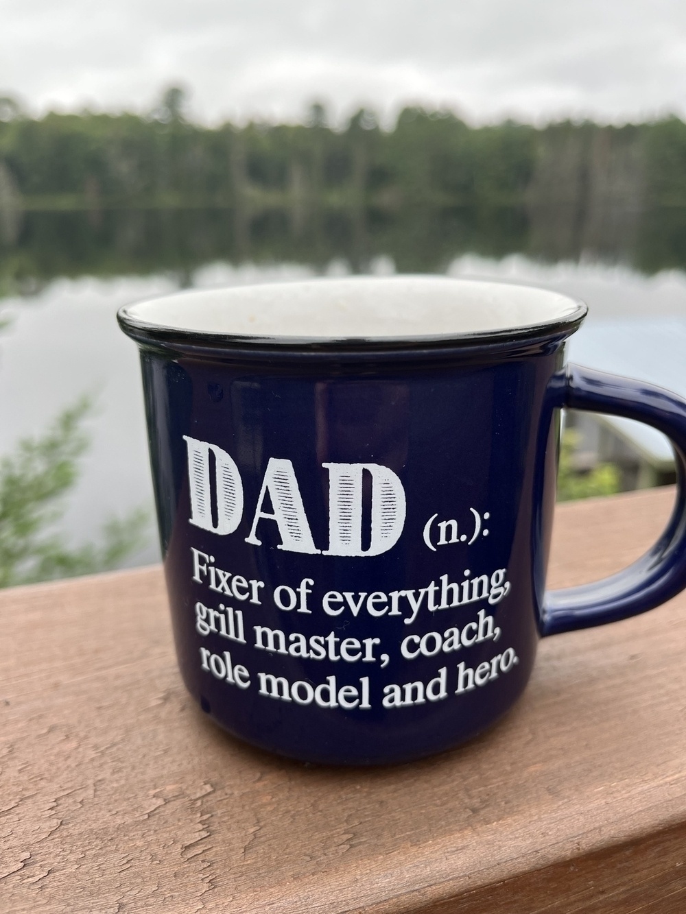 A dark blue mug with text describing the various roles of a dad is placed on a wooden surface with a serene lake and forest background.