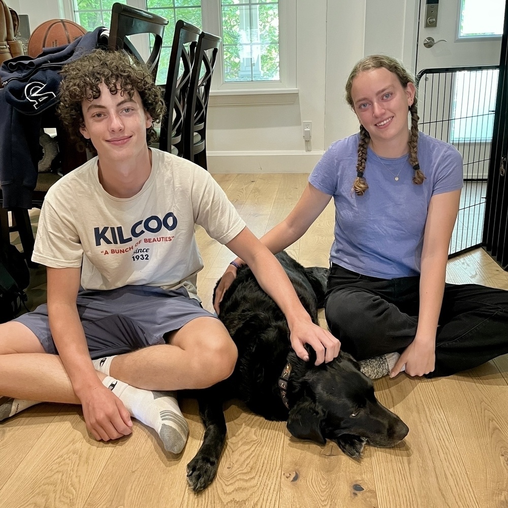 Two people are sitting on the floor with a black dog between them.