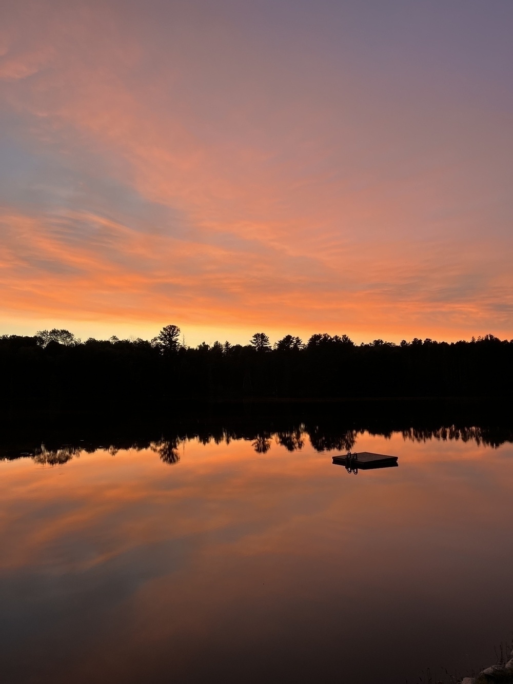 A tranquil lake reflects a vibrant sunset sky with silhouetted trees and a small dock.