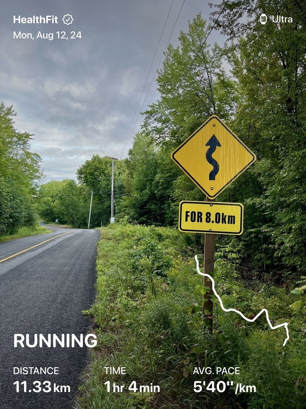 A winding road sign along a lush, green forested pathway indicates curves for the next 8.0 kilometers, with running stats overlayed on the image.