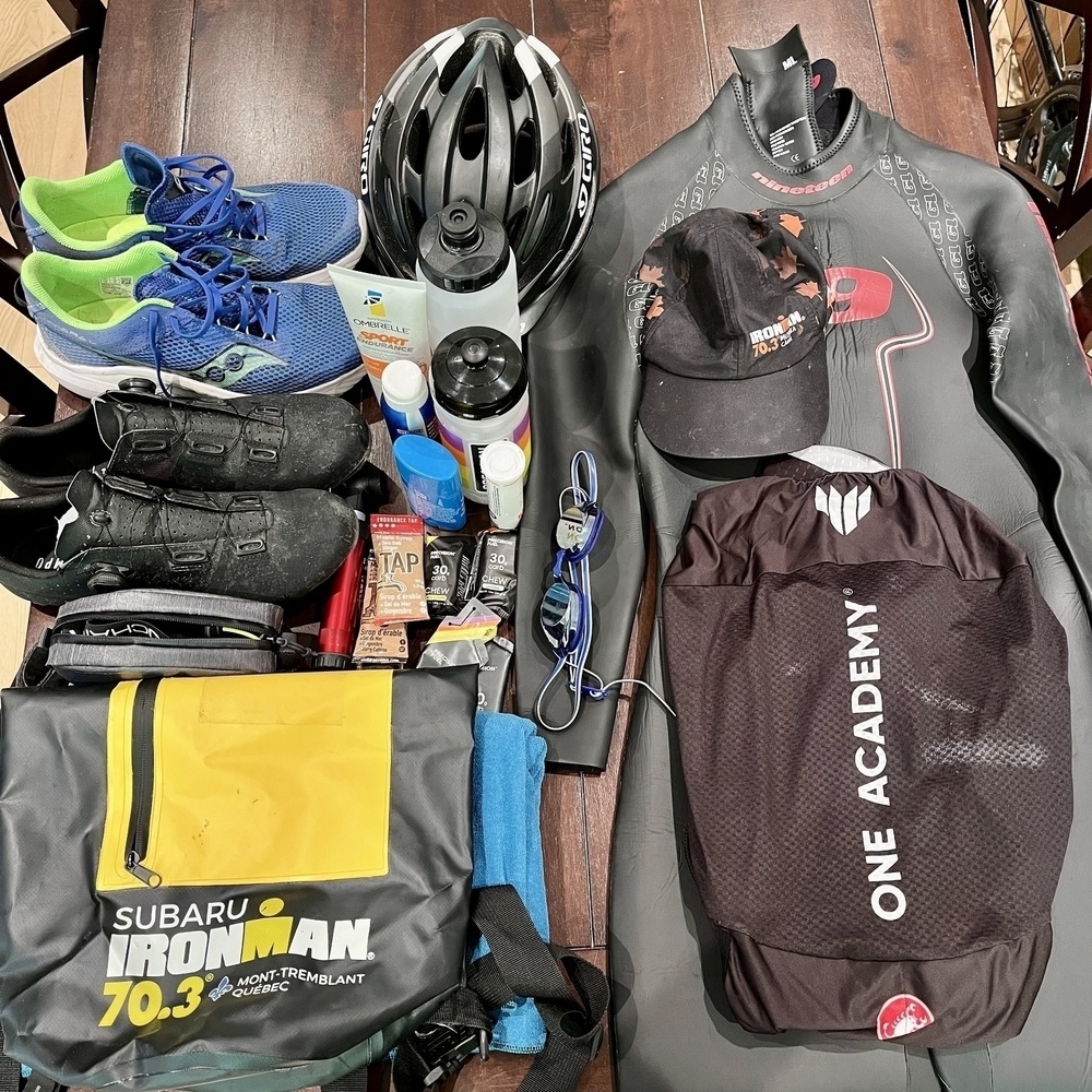 Auto-generated description: A collection of triathlon gear is neatly arranged, including running shoes, a helmet, wetsuit, various nutrition and personal care items, and branded bags.