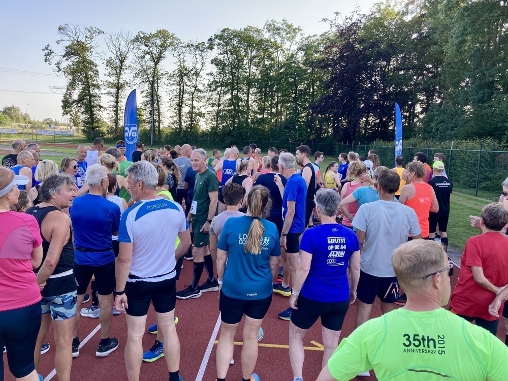 A group of people wearing athletic clothing are gathered outdoors on a track surrounded by trees and blue flags.