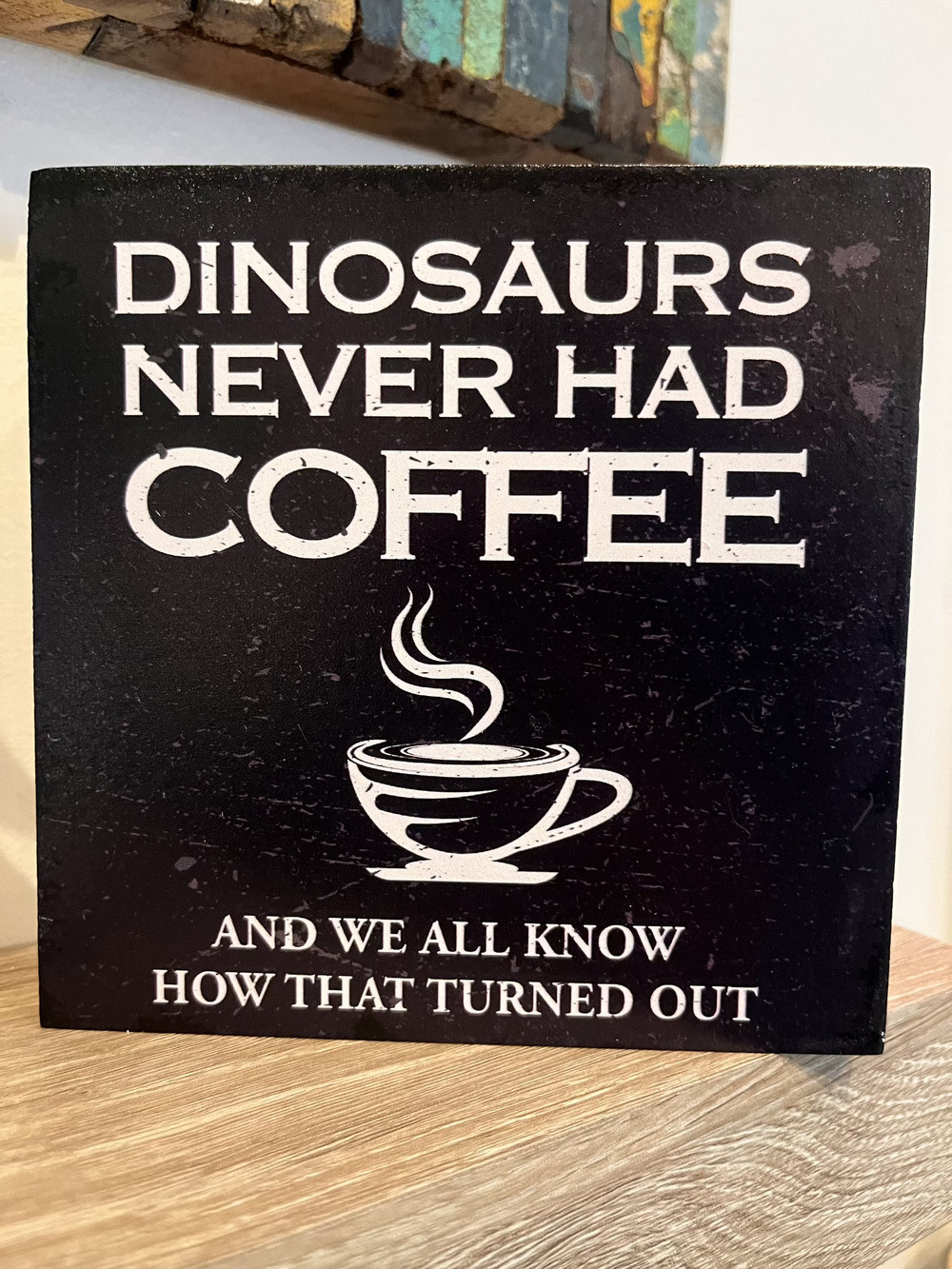 A sign on a square piece of wood on a shelf. The sign says DINOSAURS NEVER HAD COFFEE. Then a drawing of a cup of coffee, followed by, AND WE ALL KNOW HOW THAT TURNED OUT.