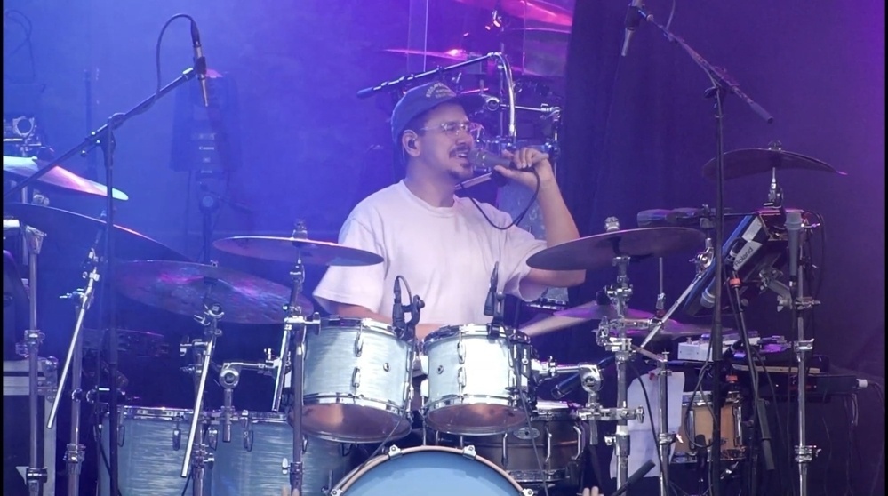 A drummer is sitting at a drum set, wearing a cap and glasses, and drinking from a bottle on stage.