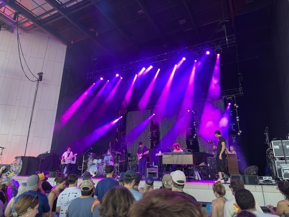 A band performs on a stage with vibrant purple lighting while an audience watches.