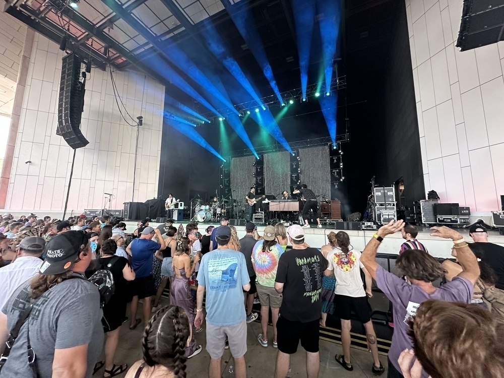 A crowd of people are watching a band perform on an indoor stage with colorful lights.