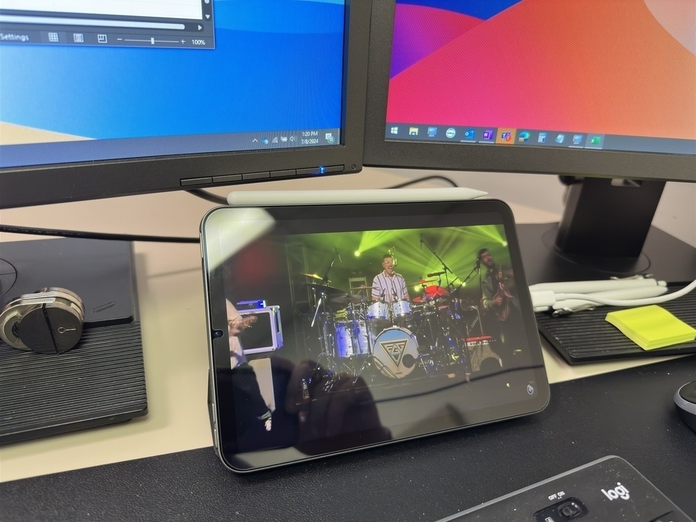 A tablet displaying a live music performance is set up in front of a dual-monitor workstation.