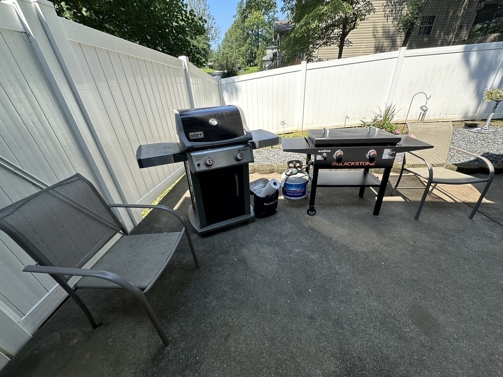A backyard patio setting includes a gas grill, a griddle, a propane tank, a metal chair, and a white fence.