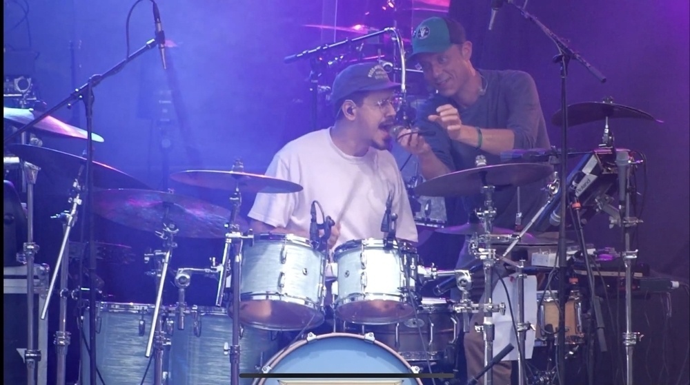 Two musicians performing on stage, one playing the drums and the other adjusting a microphone.