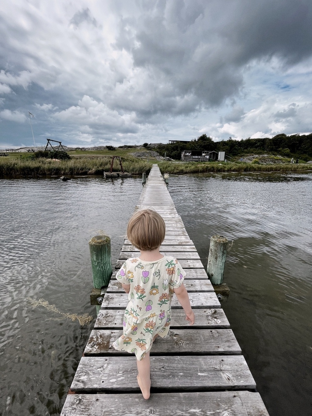 En pojke, i klänning, som promenerar på en brygga. Mot land. Bortom horisonten tornar mörka moln upp sig. 