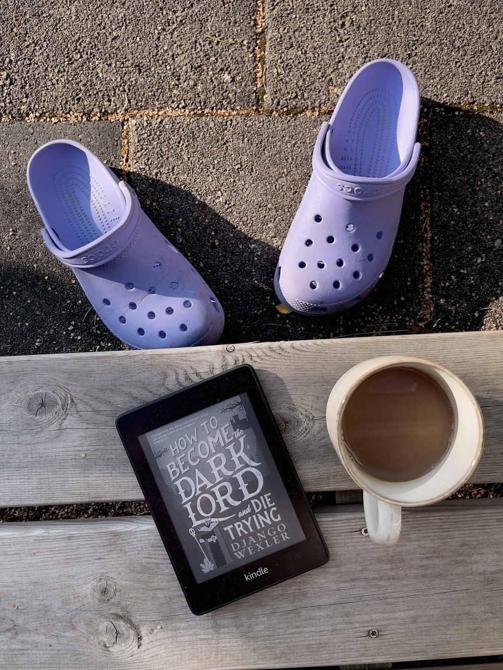 En Kindle som visar boken ”How to become the dark lord and die trying”, en kopp kaffe och ett par Crocs. 