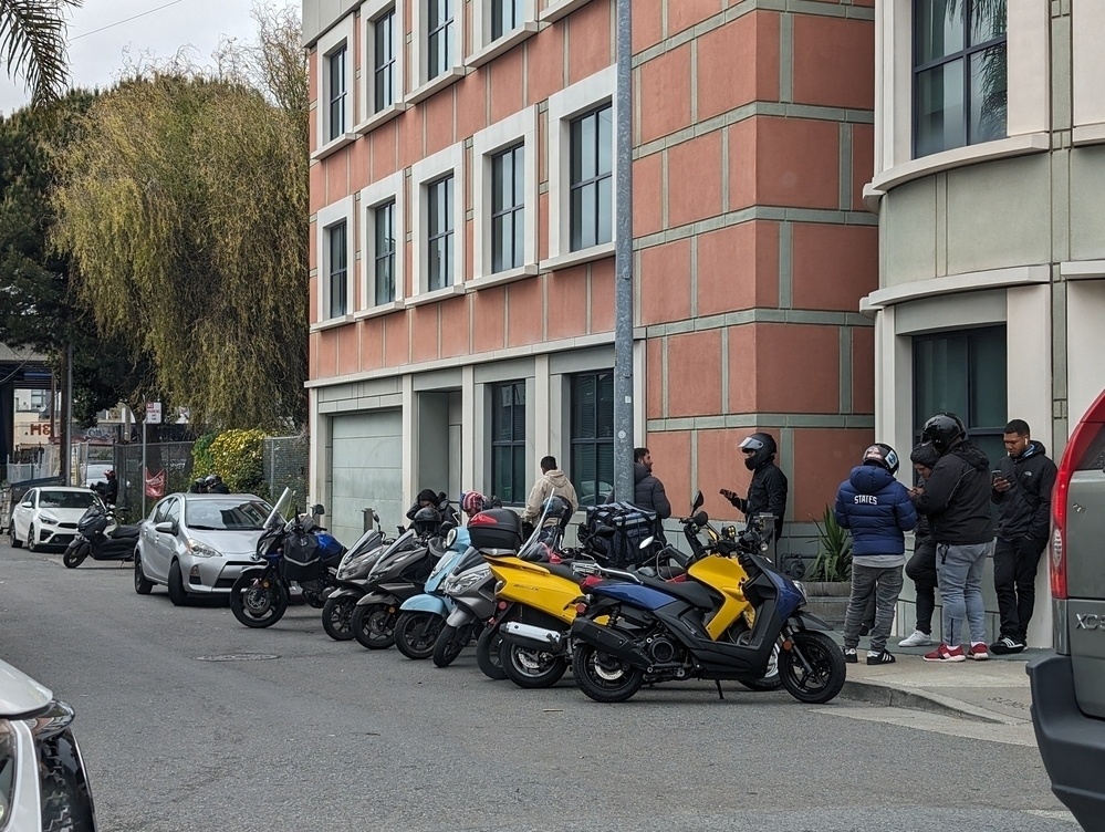 At least a half dozen electric scooters lineup along a curb in an alley with several motorcycle helmeted riders looking at phones and waiting for calls