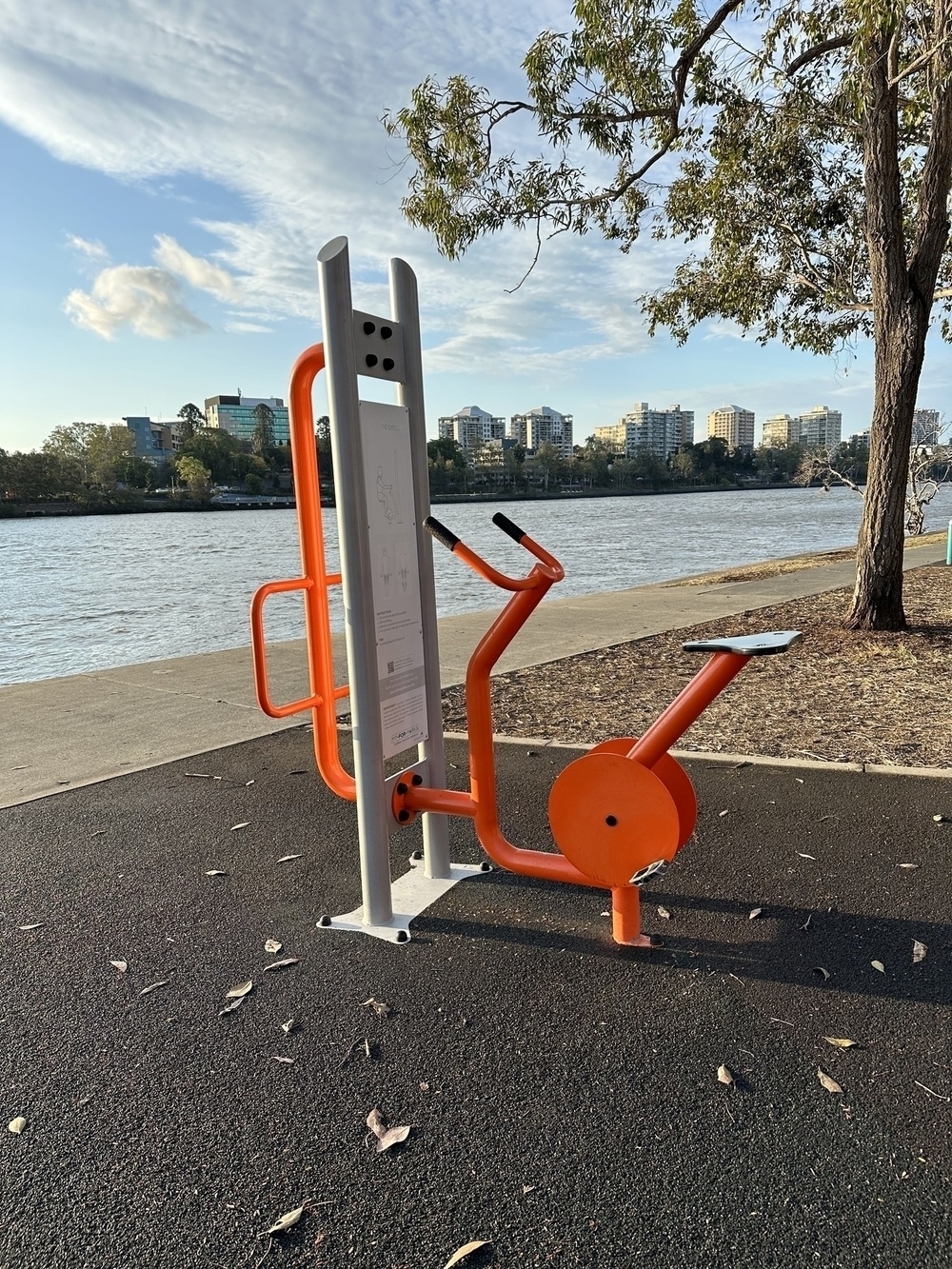 A rudimentary exercise bike made mainly from orange coloured metal pipes. Has a seat, handlebars and pedals but no actual wheels. There is grass, a tree and a river in the background.