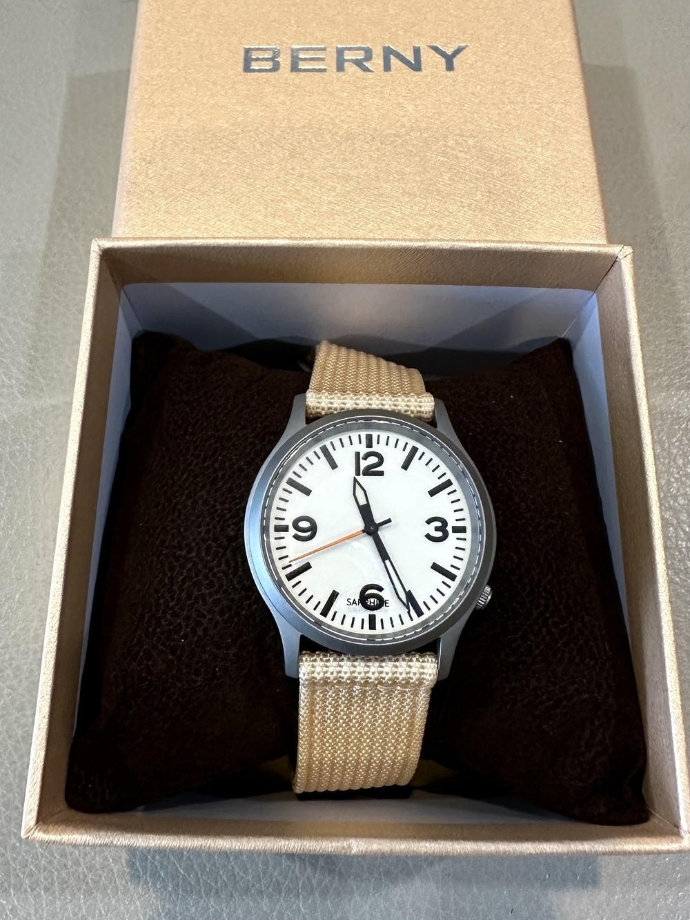 An analog watch sitting on a dark cushion inside a box. The watch has a white face with large dark 3/6/9/12 and a red second hand. The canvas strap is a cream colour.