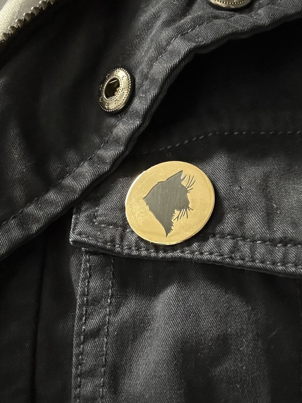 A round gold coloured badge with a black silhouette of a cat head side on. The badge is pinned to a dark jacket.