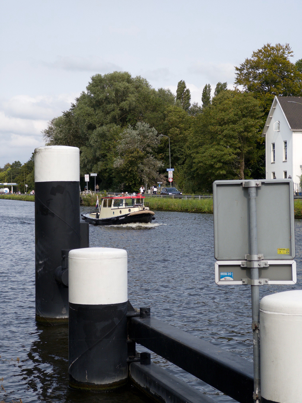 Auto-generated description: A white and black boat is navigating a river near a white building on the shore, with large black and white bollards in the foreground and trees in the background.