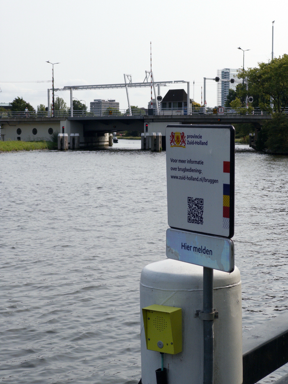 Auto-generated description: A riverside scene features a bridge, a sign with a QR code, and some urban buildings in the background.